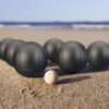 Boule-Set mit 6 Metallkugeln und Zielkugel aus Holz auf einem Sandstrand, umgeben von sanften Wellen des Ozeans.