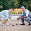 Zwei Personen spielen Boule in einem Park, eine Frau mit grauer Jacke und ein älterer Mann in grauem Overall. Sie knien und beobachten die Position der Metallkugeln auf einem Boulespielplatz mit natürlichem, grünem Hintergrund.