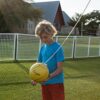 Junge spielt mit gelbem Ball im Freien, umgeben von Rasen und einem Tetherball-Netz, ideal für Garten oder Strandaktivitäten.