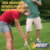Aktive Familie beim Spielen mit einem Crocket Set auf einer grünen Wiese im Freien. Die Szene zeigt eine Frau mit einem langen Schläger und einen Mann, der die Aktivität beobachtet, sowie bunte Hindernisse im Vordergrund.