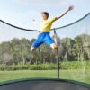 Junge springt mit Freude auf einem runden Gartentrampolin, das mit einem Sicherheitsnetz ausgestattet ist, vor einer grünen Landschaft.