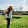 Ein fröhliches Kind springt mit einem strahlenden Lächeln auf einem roten, runden Trampolin inmitten einer grünen Wiese, umgeben von Sicherheitsnetzen und einer idyllischen Landschaft.