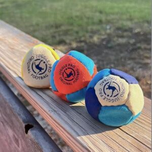 Drei bunte Juggle Pro Footbags aus Ultra Wildleder auf einer Holzbank, ideal für Freestyle und Gruppenspiele mit Sandfüllung für verbesserte Stabilität.