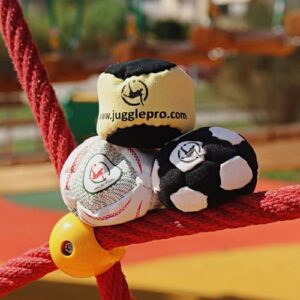 Drei Juggle Pro Footbags in verschiedenen Farben auf einem roten Seil in einer Spielplatzumgebung, geeignet für Freestyle und Gruppenspiele.