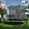 Großes rundes Trampolin 'Ultimate Pro' im Kürbis-Design, umgeben von Sicherheitsnetz und Leiter, im gepflegten Garten mit zwei Personen, die auf das Trampolin schauen.