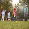 Gruppenszene im Freien mit mehreren Personen, die Krocket spielen. Eine Frau mit rotem Oberteil schlägt einen gelben Ball mit einem Krocket-Schläger auf dem grünen Rasen. Im Hintergrund sind das Set und kleinere Tore zu sehen, umgeben von einer grünen Hecke.