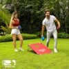 Ein Paar spielt Cornhole im Freien, mit einem roten Spielbrett und blauen sowie roten Spielbeuteln. Die Freude an dieser beliebten amerikanischen Freizeitaktivität ist deutlich sichtbar vor einer üppigen grünen Landschaft.