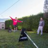 Mädchen balanciert auf einer Slackline, unterstützt von zwei robusten Stützen, umgeben von einem grünen Rasen und sportlich aussehenden Beobachtern an einem sonnigen Tag.