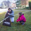 Vater und Tochter beim Aufbau einer Slackline im Garten mit Holzstütze und drei Höhenverstellungen für unterschiedliche Erfahrungsgrade.