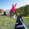 Ein Mädchen balanciert auf einer Slackline im Freien, umgeben von grüner Rasenfläche und Bäumen, während es das Gleichgewicht trainiert.