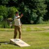 Mann wirft einen Sack auf eine Cornhole-Spielplattform im Garten, umgeben von grünen Bäumen und Gras - ideal für entspannte Freizeitaktivitäten.