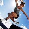 Junge Frau springt dynamisch in die Luft, während sie einen grauen Footbag in der Hand hält, umgeben von hellblauem Himmel und Sonnenlicht, was die Freude am Freestyle-Footbag verdeutlicht.